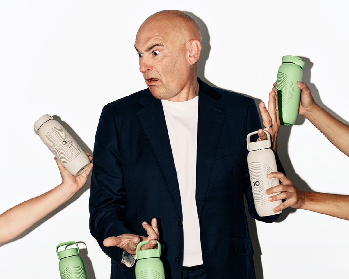 Homme en veste bleu foncé et chemise blanche entouré de mains offrant plusieurs bouteilles d'eau réutilisables de différentes couleurs.