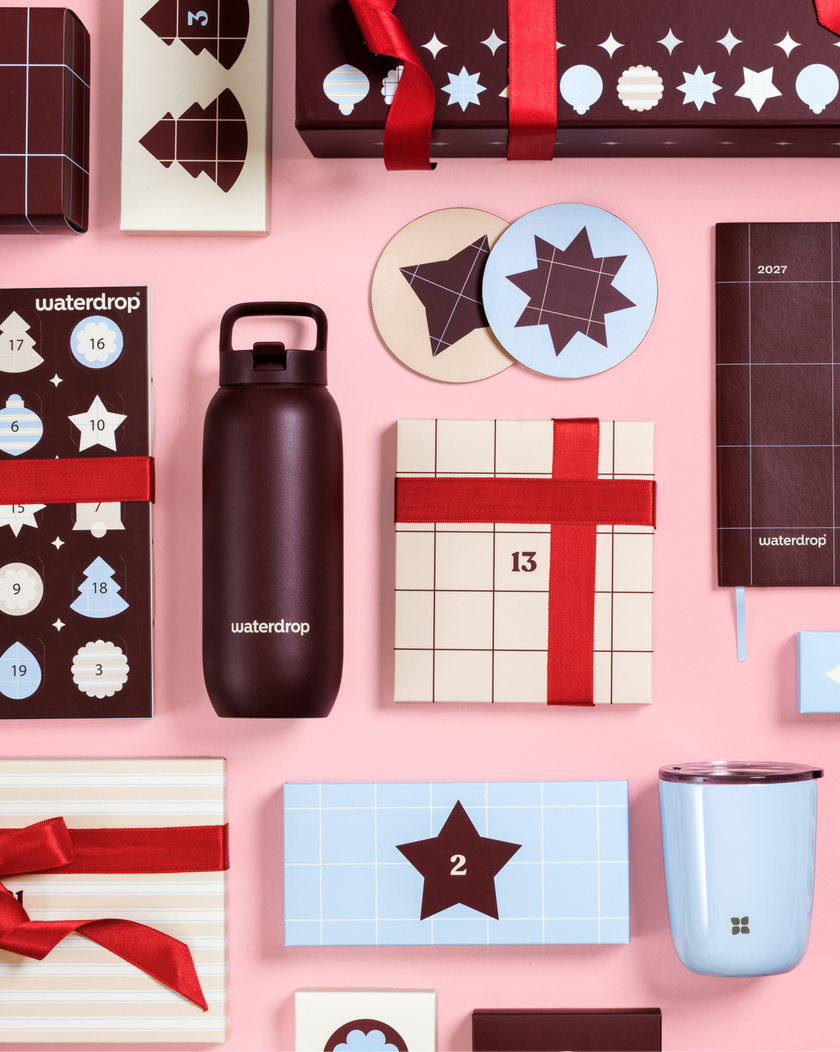 Calendriers de l'Avent et boîtes cadeaux à rubans rouges sur fond rose, avec une bouteille noire et une tasse bleu clair.