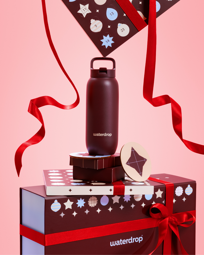 Bouteille d'eau noire avec bouchon et poignée sur des coffrets-cadeaux empilés, noués de rubans rouges sur fond rose.
