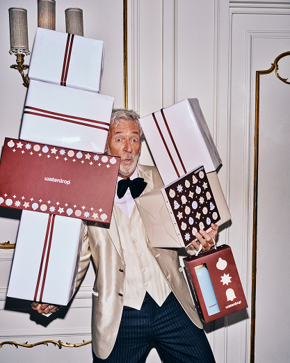 Homme en costume beige porte plusieurs cadeaux emballés à l’intérieur.