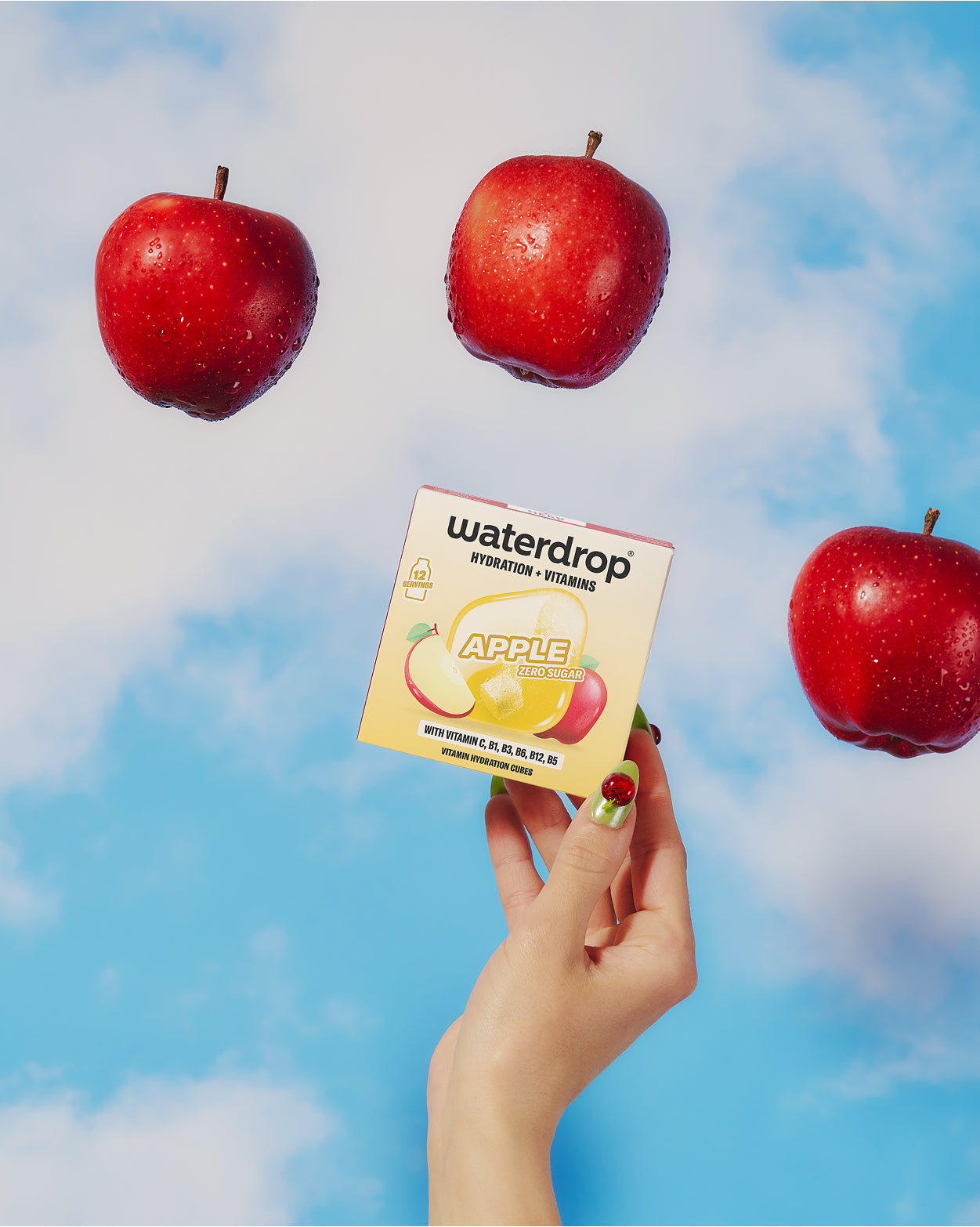 Main tenant une petite boîte de produit avec une illustration de pomme, sur un ciel bleu avec trois pommes rouges flottantes.