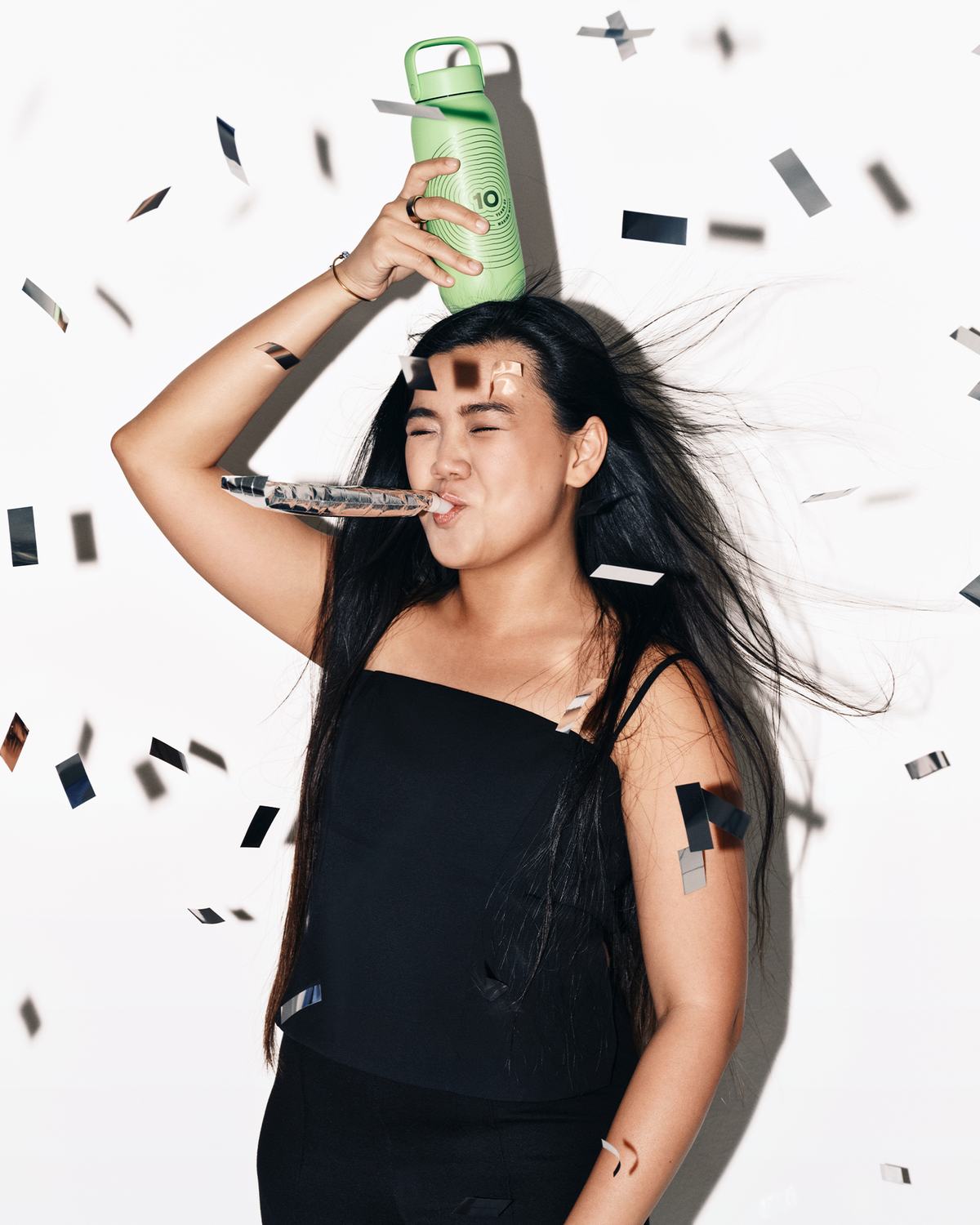 Femme en haut noir tenant une bouteille de spray verte sur la tête, des confettis tombent autour.