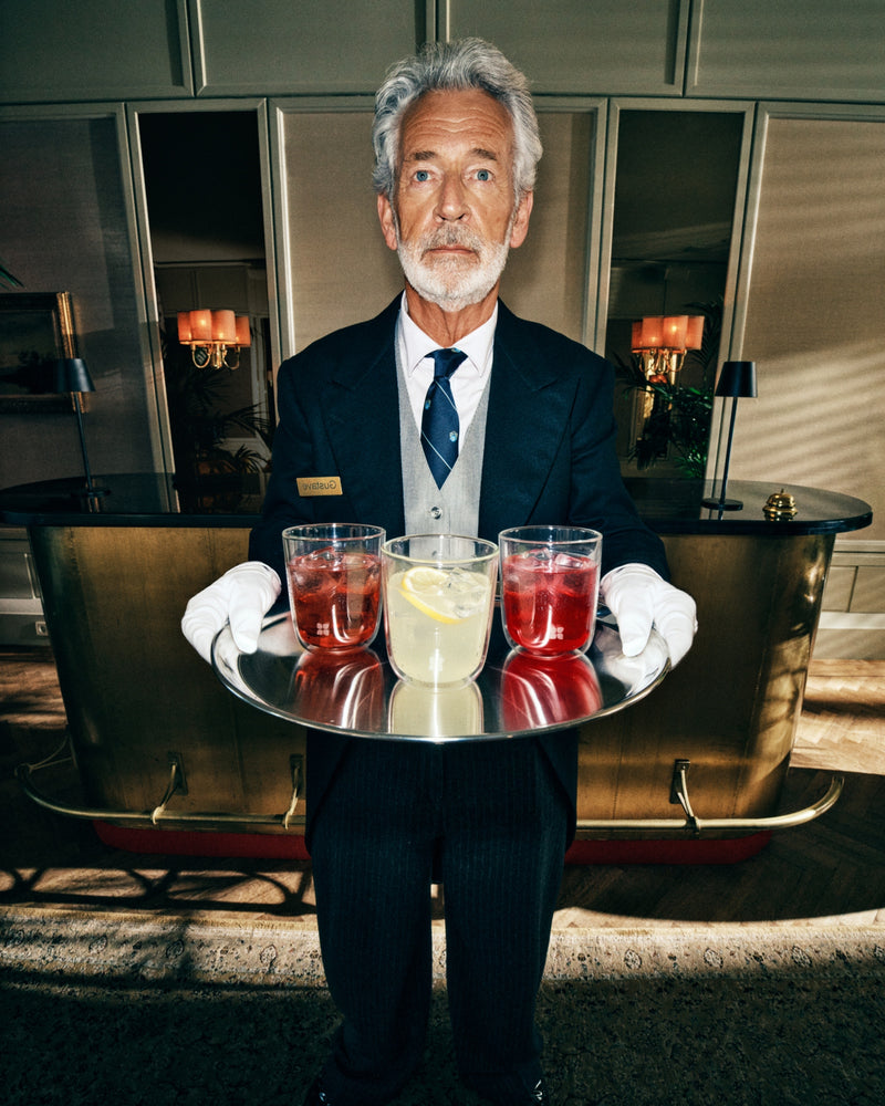 Assistant server en costume sombre présente un plateau rond avec deux boissons rouges et une boisson jaune pâle dans le hall d’un hôtel.