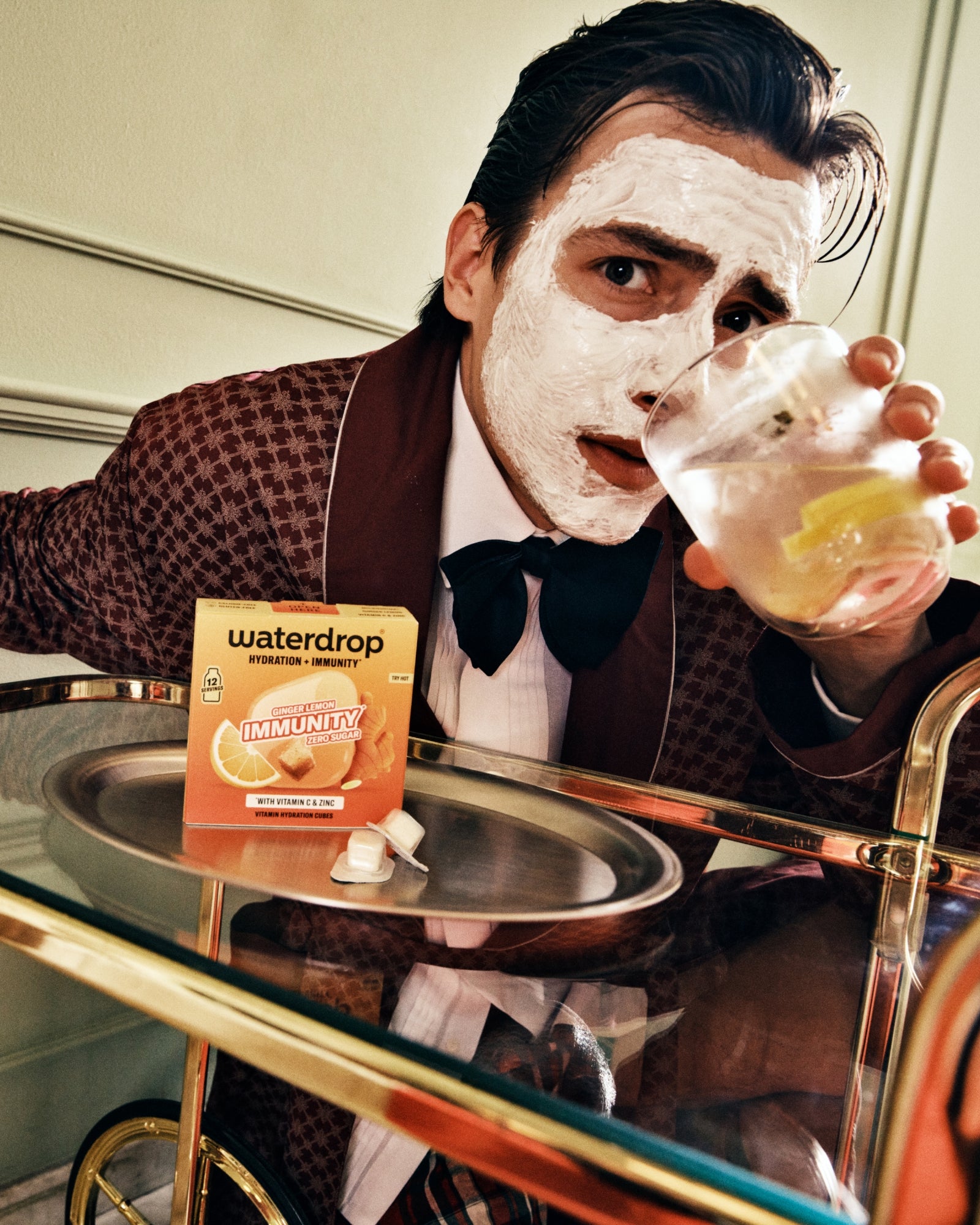 Homme en costume brun portant un masque facial et buvant dans un verre au comptoir.