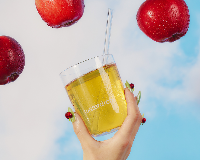 Une main tient un verre de jus jaune avec une paille, entouré de pommes rouges et d'un ciel bleu.