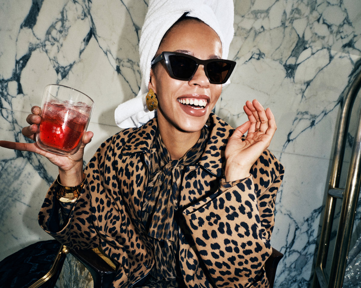 Femme portant un manteau leopard et des lunettes de soleil, avec une serviette blanche sur la tête, tenant une boisson rouge dans un verre devant un mur en marbre.