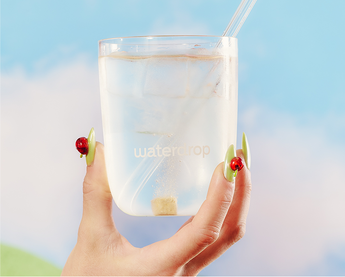 Une main tient un verre d'eau transparent avec des glaçons et une paille, sur fond de ciel bleu.