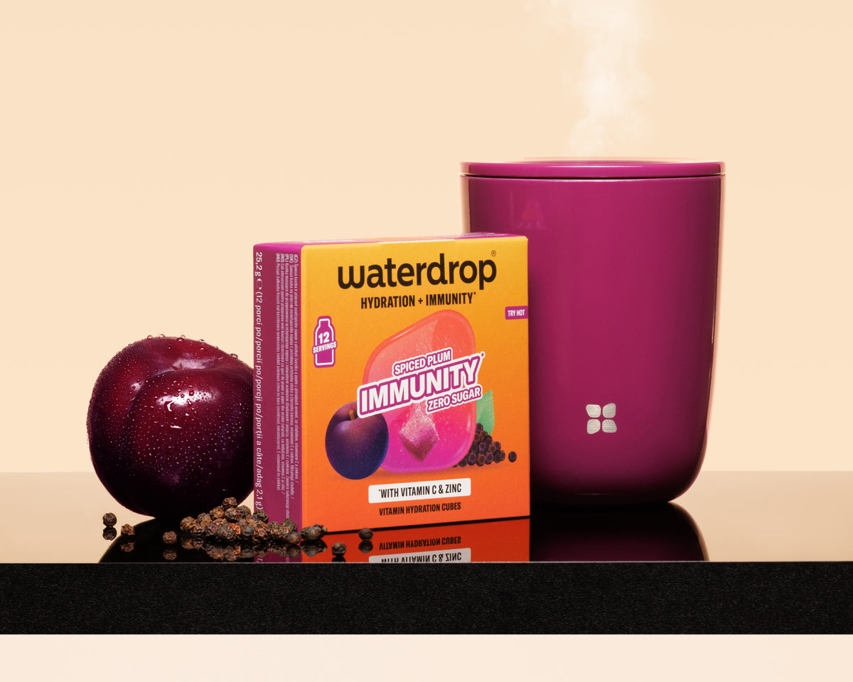 Tasse violette avec vapeur à côté d'une boîte orange Waterdrop, une prune et des graines éparpillées sur une surface réfléchissante.