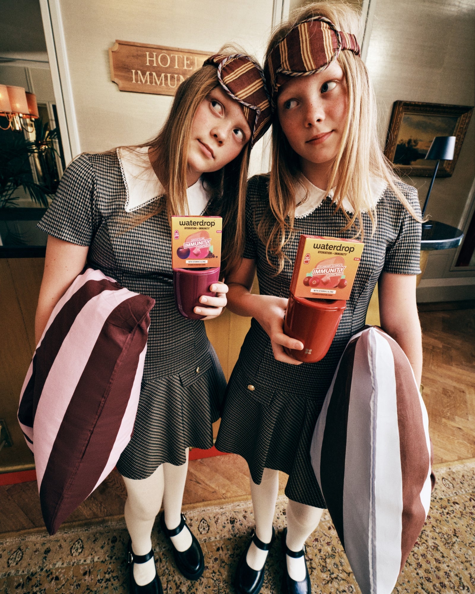 Deux filles en robes à carreaux assorties et serre-têtes rayés se tiennent côte à côte dans le hall d'un hôtel, chacune tenant un sachet de jus.